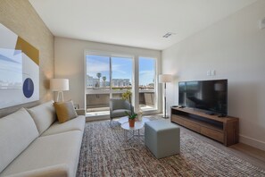 A modern living room with a sofa, coffee table, and TV, large windows with a city view, and a balcony with a chair.
