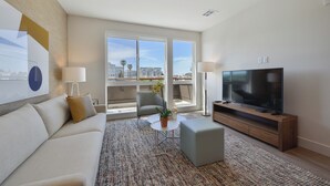 Una sala moderna con sofá, mesa de centro y televisor, ventanas amplias con vista a la ciudad y un balcón con una silla.
