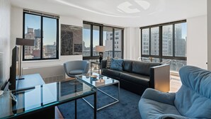 Ein modernes Wohnzimmer mit einem Glastisch, einem schwarzen Sofa, einem blauen Sessel und einem Schreibtisch mit einer Lampe. Große Fenster bieten Blick auf die Stadt.
