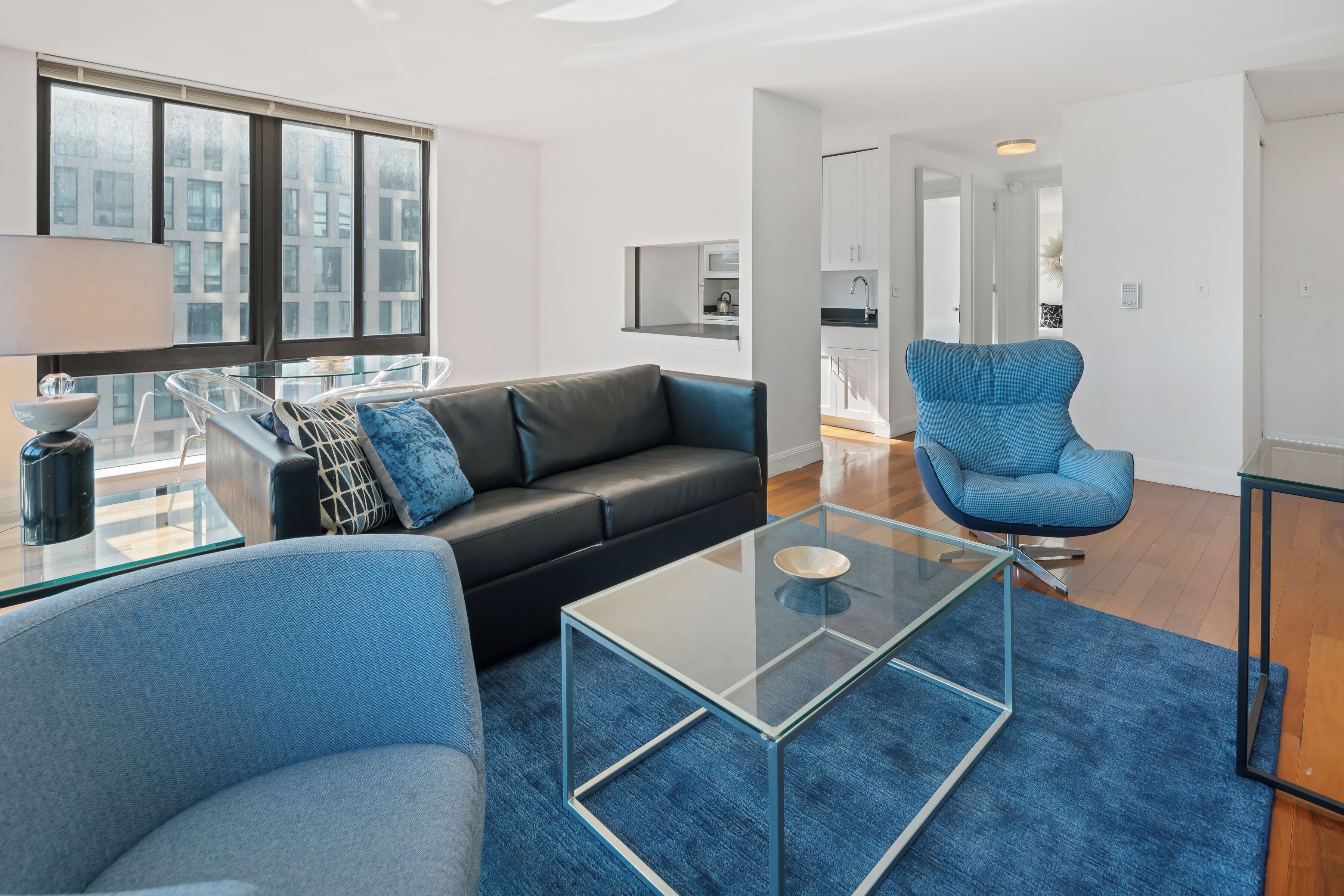 A modern living room with a glass coffee table, a black leather sofa, a blue armchair, and a glass dining table.