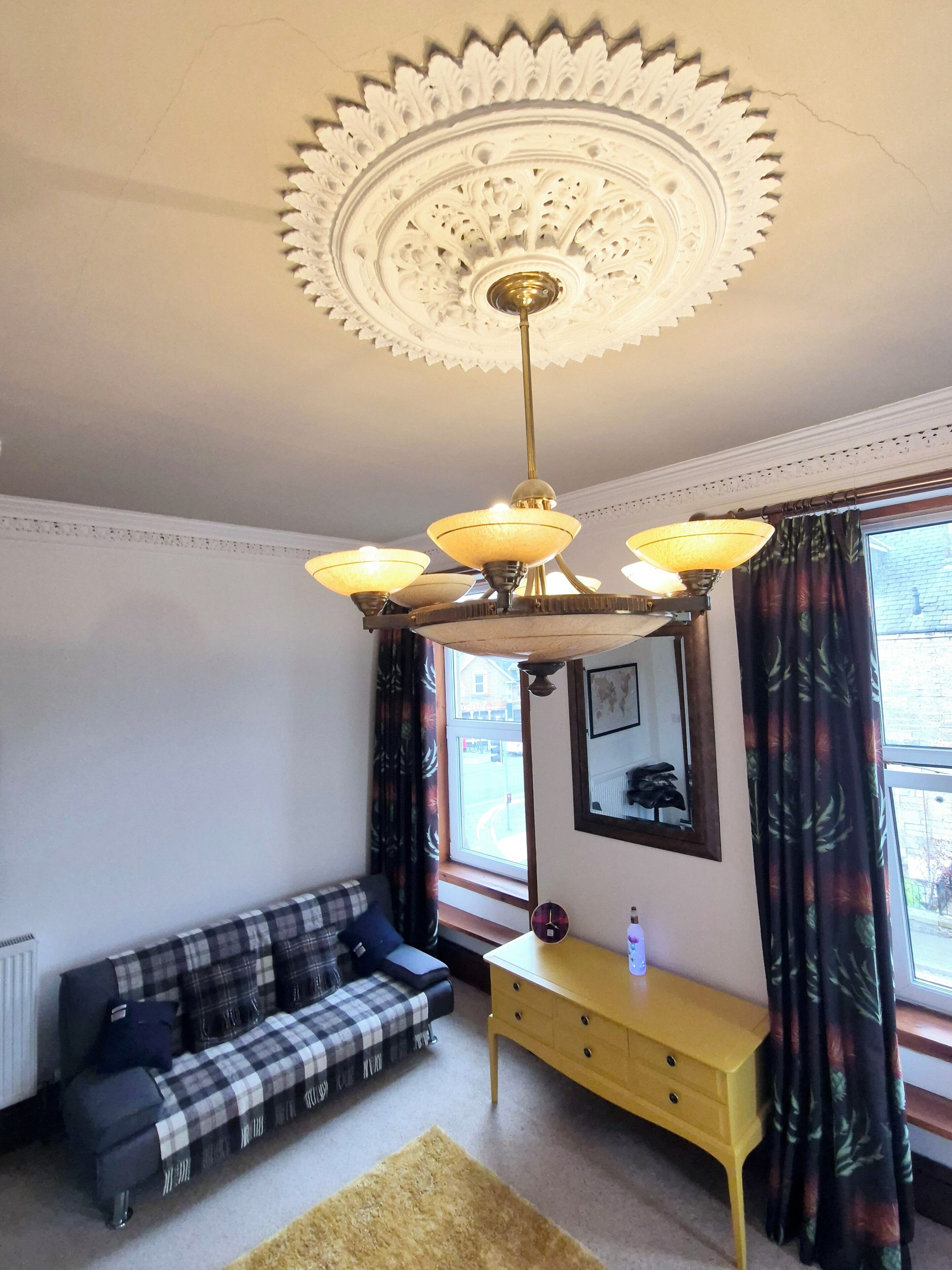 A room with a checkered sofa, a yellow dresser, a decorative ceiling, and a window with curtains.