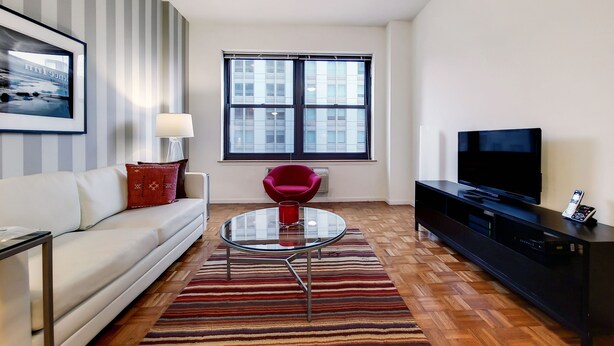 Una sala de estar moderna con un sofá blanco, una mesa de centro de vidrio, una silla roja, un mueble de entretenimiento negro y un ventanal grande.