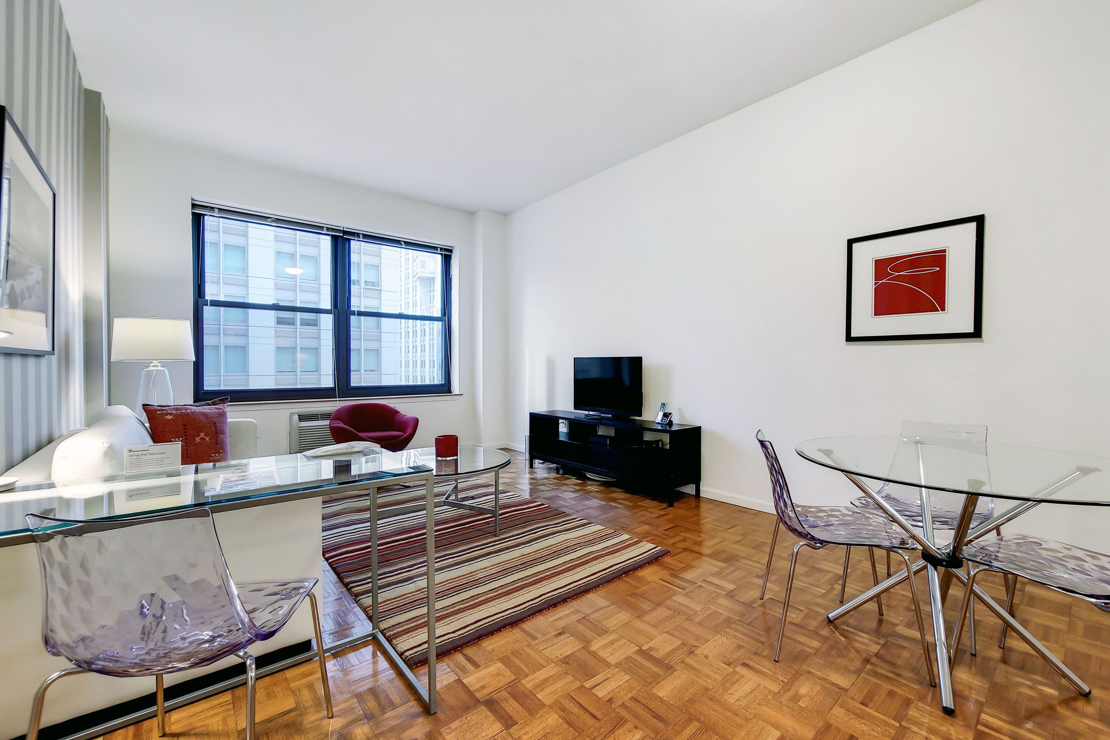A modern living room with a glass dining table, clear chairs, a sofa, a TV, and a large window.