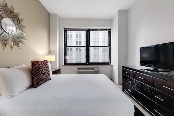 A hotel room with a large bed, a flat-screen TV, a window with blinds, a heater, and a wall-mounted mirror.