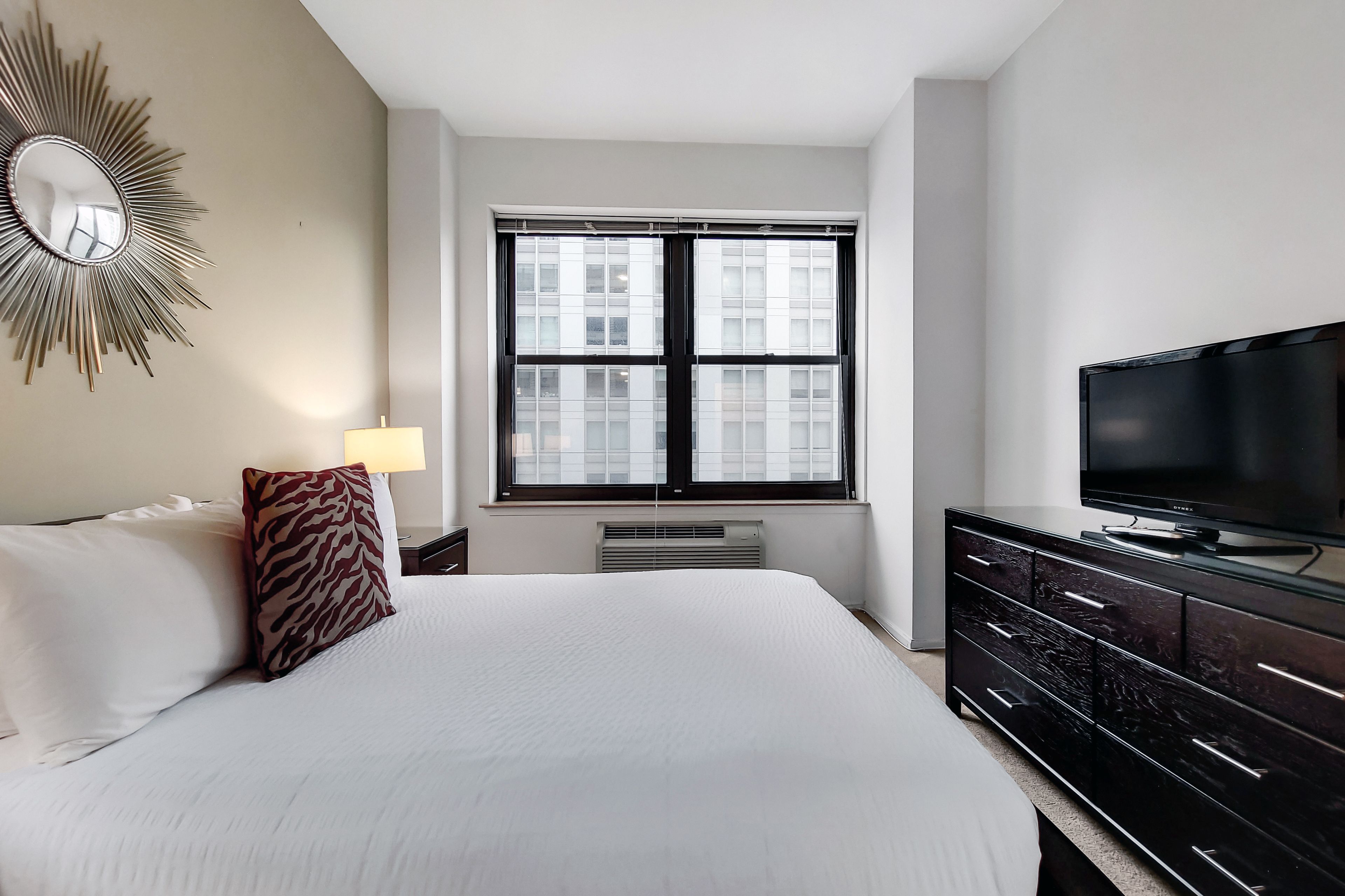 A hotel room with a large bed, a flat-screen TV, a window with blinds, a heater, and a wall-mounted mirror.