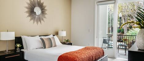 A modern bedroom with a large bed, two bedside tables, a decorative mirror, and a view of the outdoors through a sliding glass door.