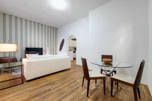 A modern living room with a white sofa, a glass coffee table, and a TV. There is a dining area with a glass table and chairs. The room has wooden flooring and white walls.