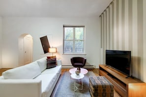 A modern living room with a white sofa, a small round table, a dark armchair, a TV, and a window with a view of trees. - Central Park Living | UWS | On-Site Laundry (New York)