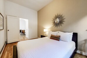 A bedroom with a large bed, a wall-mounted mirror, a door leading to another room, and a small table with a lamp.