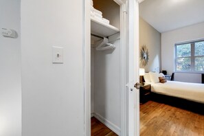 A bedroom with a wooden floor, a bed, a nightstand, a lamp, and a window with a view of trees. - Central Park Living | UWS | On-Site Laundry (New York)