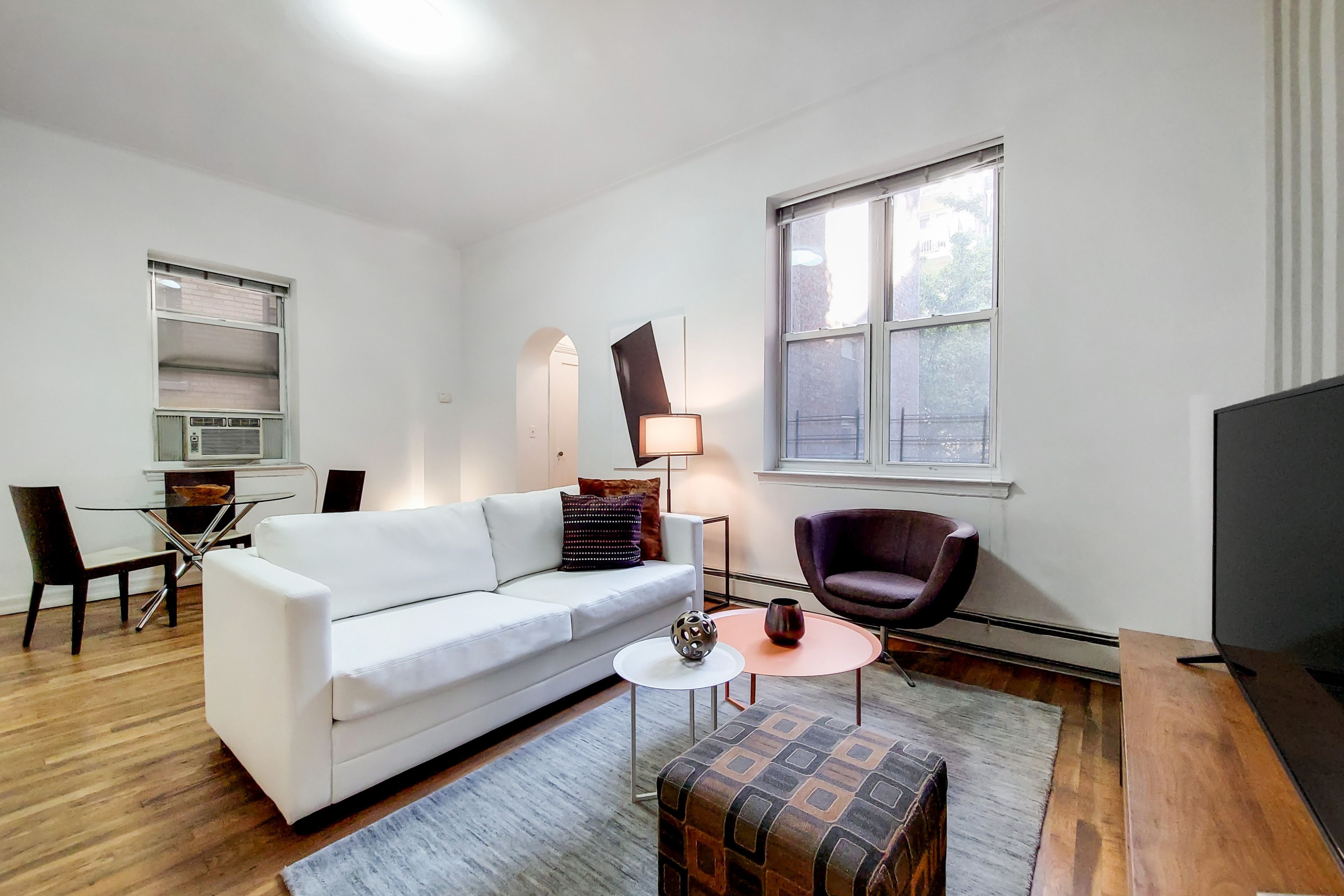 A modern living room with a white sofa, a round coffee table, a patterned ottoman, a purple armchair, and a TV.