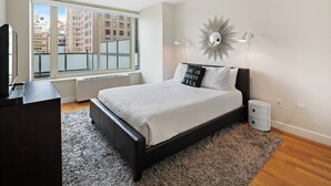 Quarto moderno com uma janela ampla, cama com edredom branco, cabeceira preta e um objeto decorativo na parede.