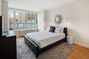 Ein modernes Schlafzimmer mit großem Fenster, einem Bett mit weißer Bettdecke, einem schwarzen Kopfteil und einem dekorativen Wandbild.