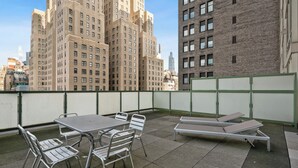 Um terraço no telhado com mesas e cadeiras de metal, um longo banco e vista para prédios altos ao fundo.