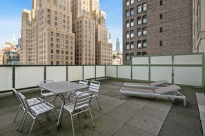 Eine Dachterrasse mit Metalltischen und -stühlen, einer langen Bank und Blick auf hohe Gebäude im Hintergrund.