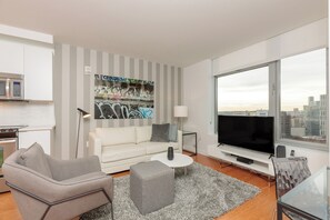 Un salon moderne avec un canapé, un pouf et un téléviseur, un coin cuisine équipé d’un four à micro-ondes, et une grande fenêtre offrant une vue sur la ville.