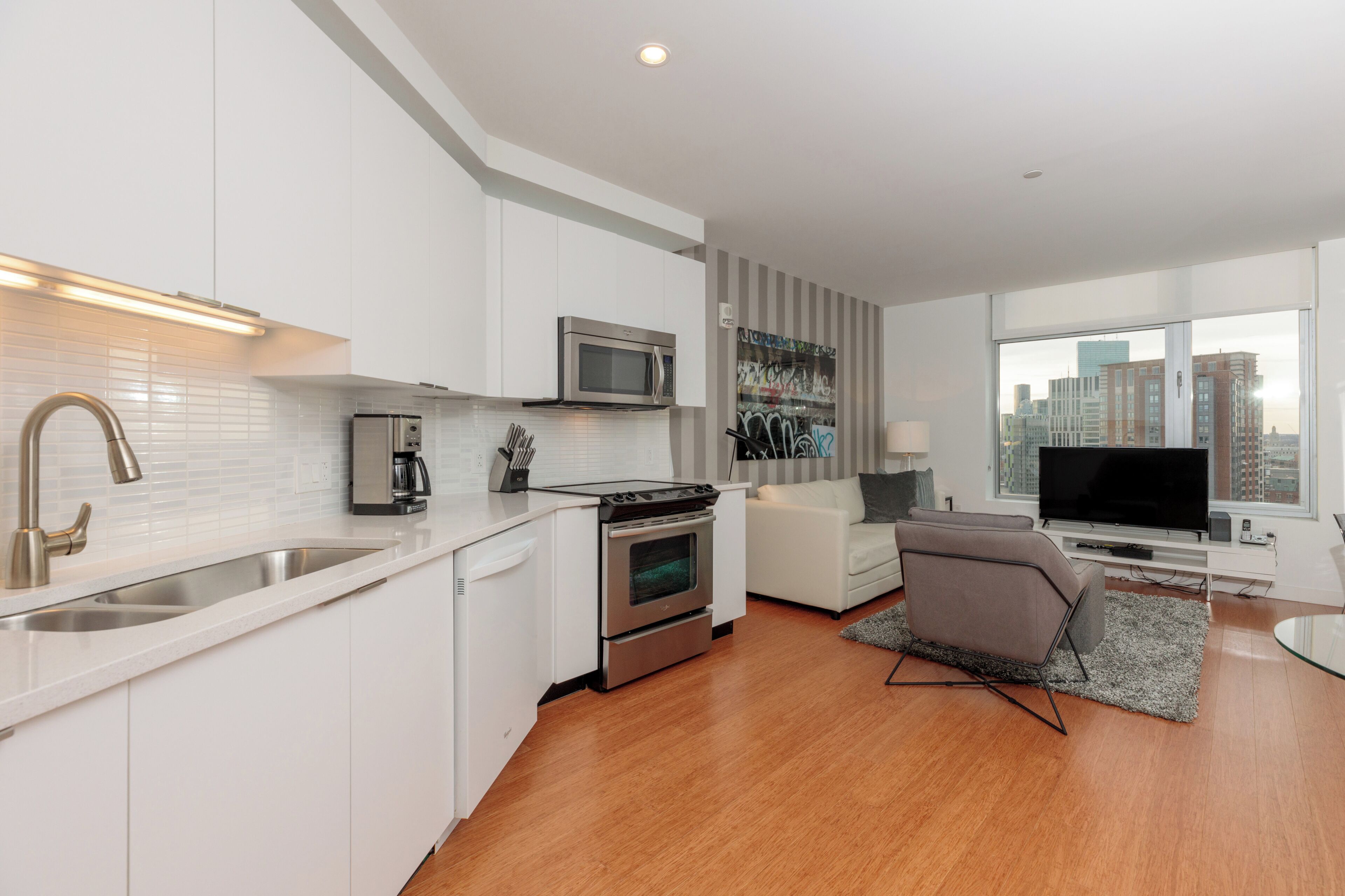 A modern kitchen with white cabinetry, a stainless steel sink, a microwave, and an oven. There is a dining area with a white sofa and a gray rug. A flat-screen TV is mounted on the wall.