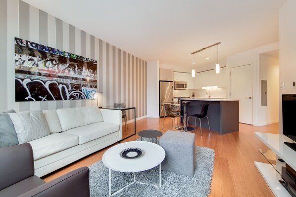 A modern living room with a sofa, a coffee table, and a rug. There is a kitchen area with a microwave, refrigerator, and dining table.