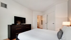 A modern bedroom with a large bed, a dark dresser, a flat-screen TV, and a bedside lamp. - Modern 1 Bedroom in Downtown Boston (Boston)