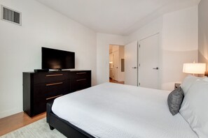A modern bedroom with a large bed, a dark dresser, a flat-screen TV, and a bedside lamp. - Modern 1 Bedroom in Downtown Boston (Boston)