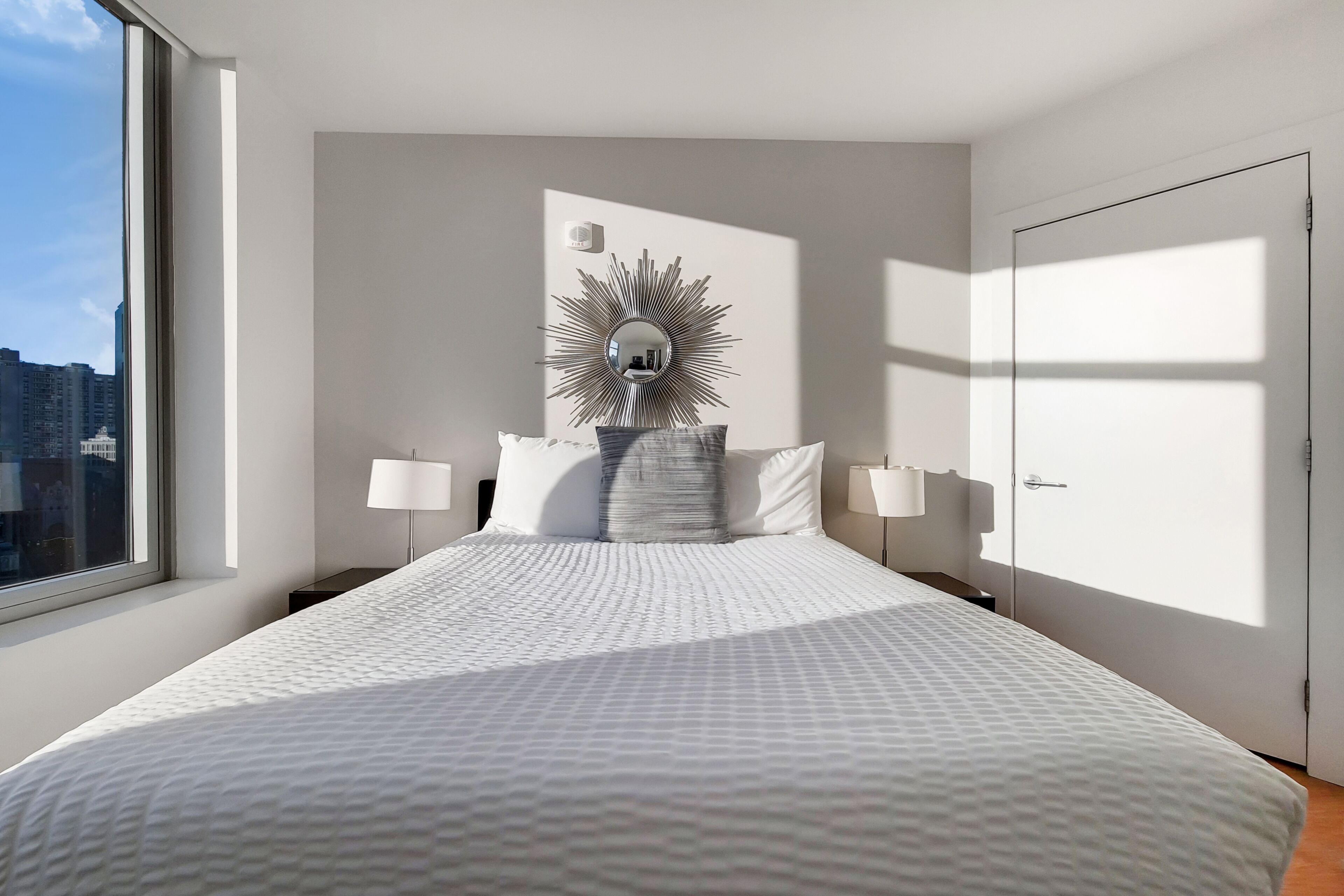 A modern hotel room with a large bed, a sunburst mirror above the headboard, and a city view through the window.
