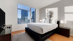 A modern bedroom with a large window, a bed with white bedding, a side table with a lamp, and a decorative wall piece.