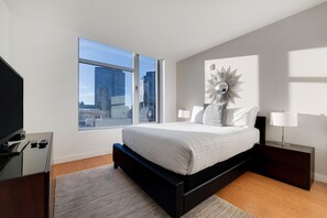 Quarto moderno com uma janela ampla, cama com roupa de cama branca, mesa de cabeceira com abajur e um objeto decorativo na parede.