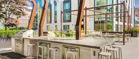 An outdoor bar area with a grill, bar stools, and a view of a modern building with large windows.