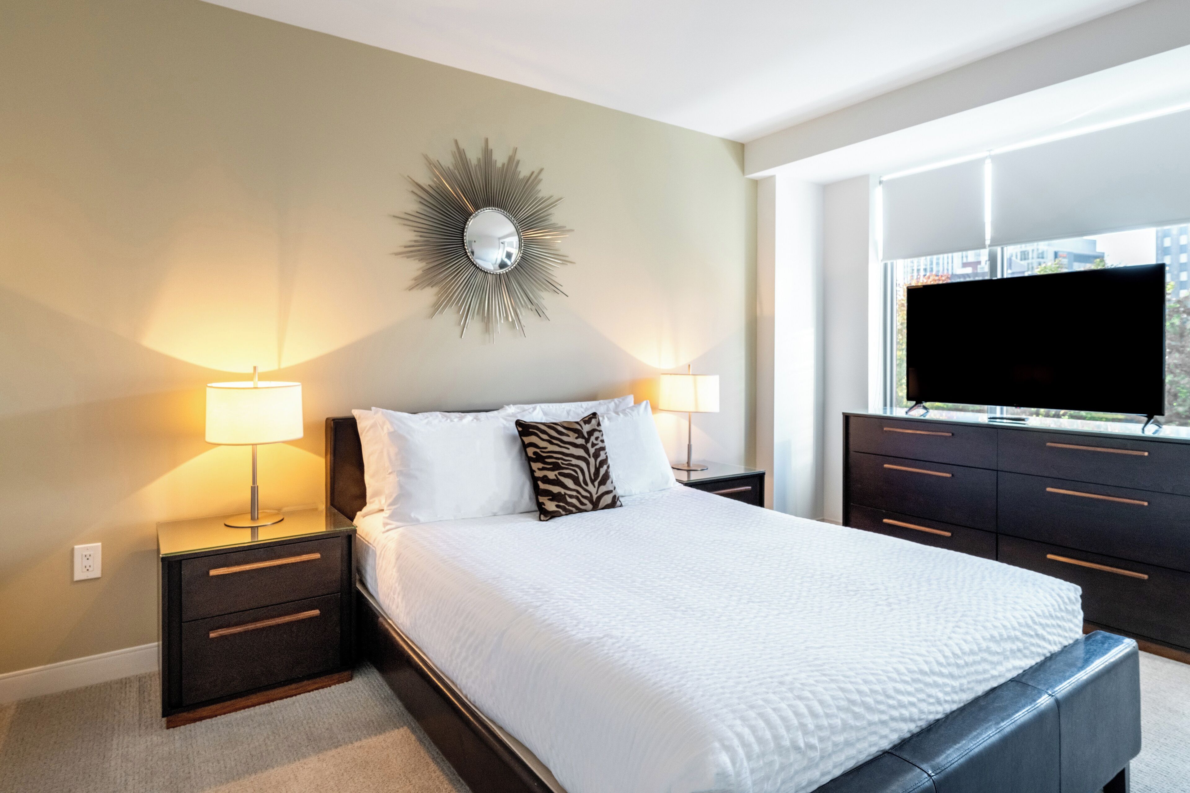 A hotel room with a large bed, two bedside tables with lamps, a wall-mounted mirror, a flat-screen TV, and a window with a city view.
