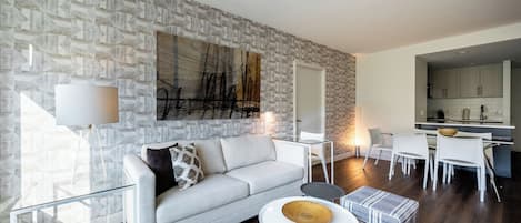 A modern living room with a grey sofa, a coffee table, and a dining area with white chairs and a kitchen in the background.