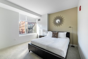 Une chambre moderne avec une grande fenêtre, un lit recouvert de linge de lit blanc, un miroir décoratif sur le mur et une lampe sur pied.