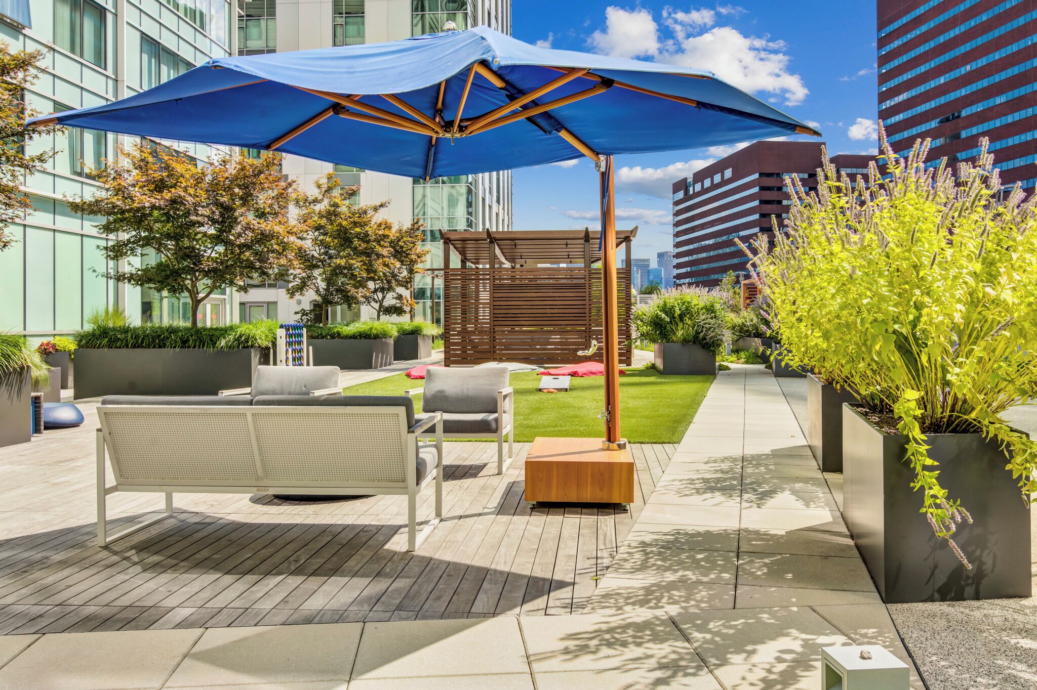 Een terras met een blauwe parasol, een houten pergola en moderne gebouwen op de achtergrond.