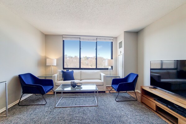 A modern living room with a sofa, two blue chairs, a coffee table, a TV, and large windows with a city view.