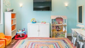 Um espaço de recreação infantil com televisão fixada na parede, um cercadinho, uma casa de bonecas e um tapete colorido.