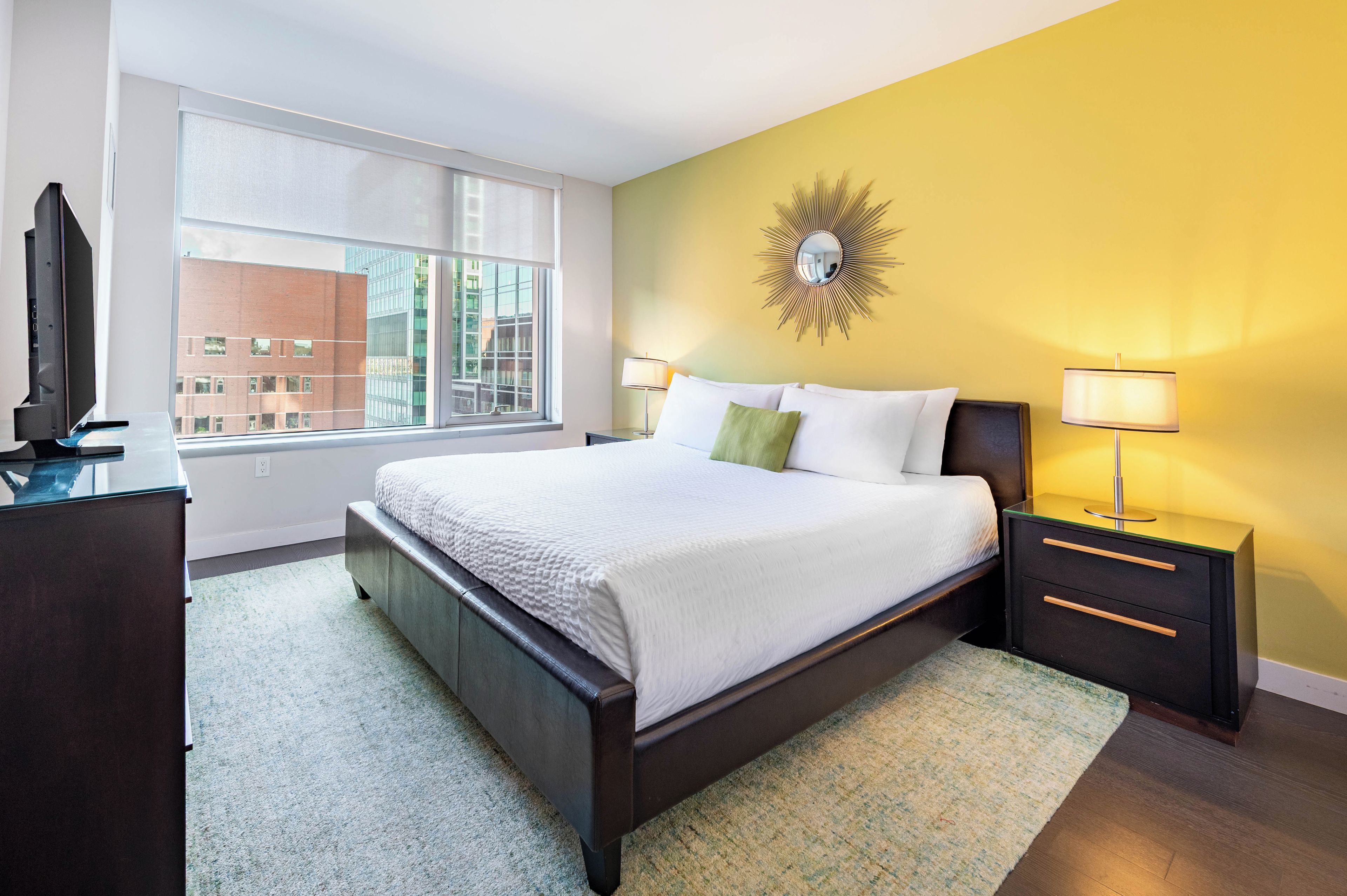 A modern hotel room with a large bed, a television, a window with blinds, a nightstand with a lamp, and a decorative wall piece.