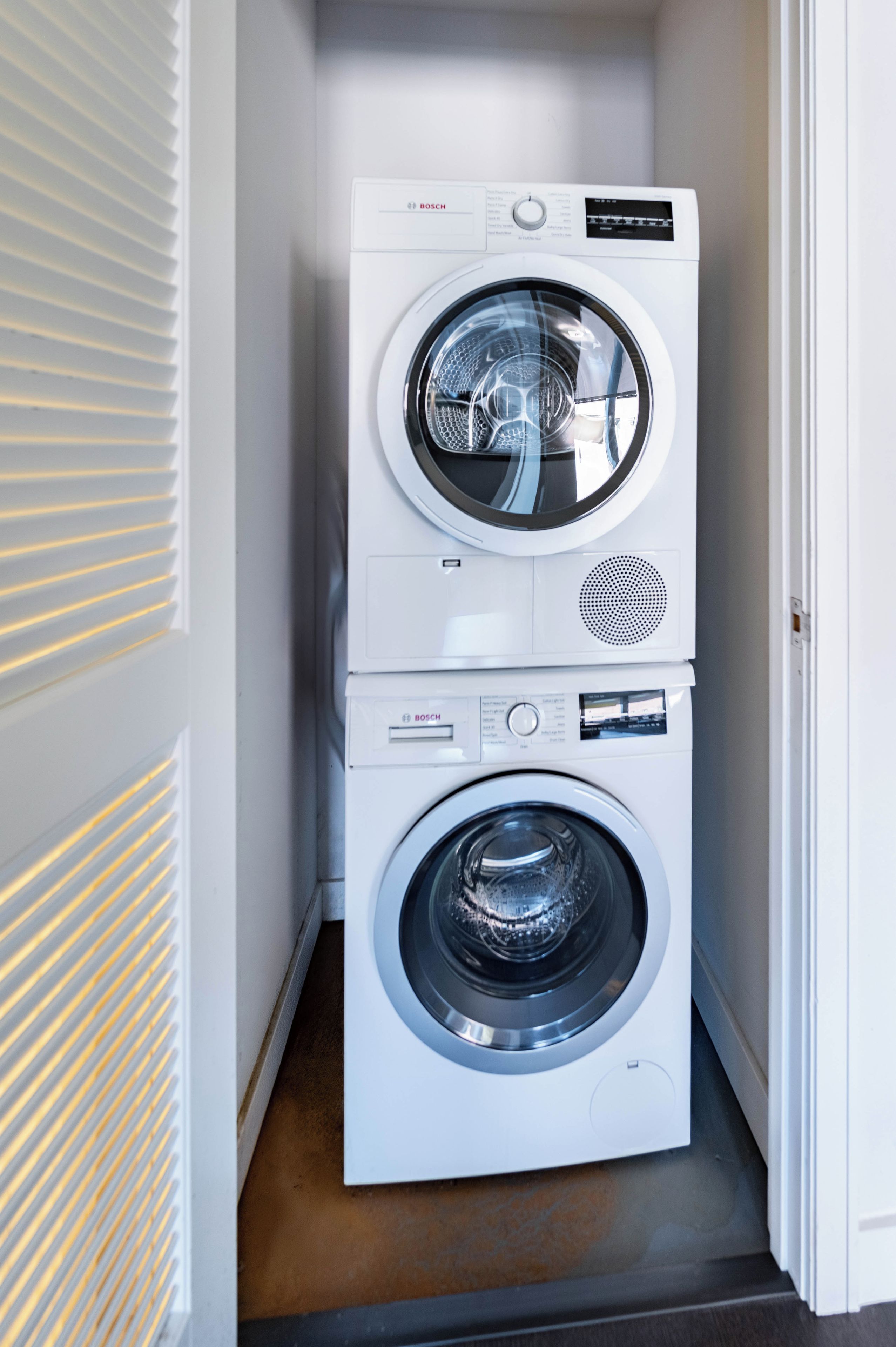 A stacked Bosch washing machine in a laundry room with a door and blinds.