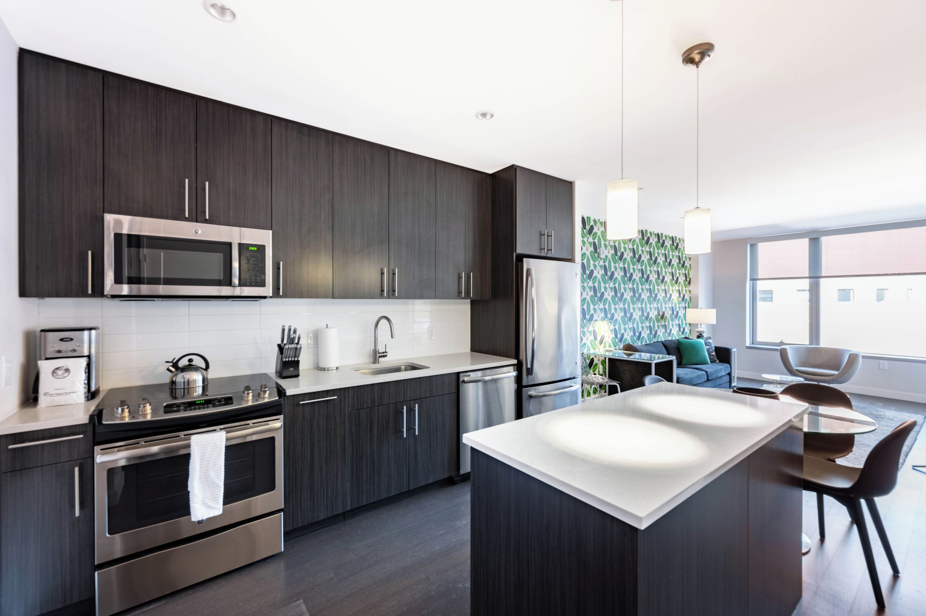A modern kitchen with dark cabinetry, stainless steel appliances, a central island, and a view of the living area.