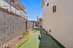 A narrow pathway with a small garden area, a blue flag, and a building with a white exterior and a brick wall on one side.