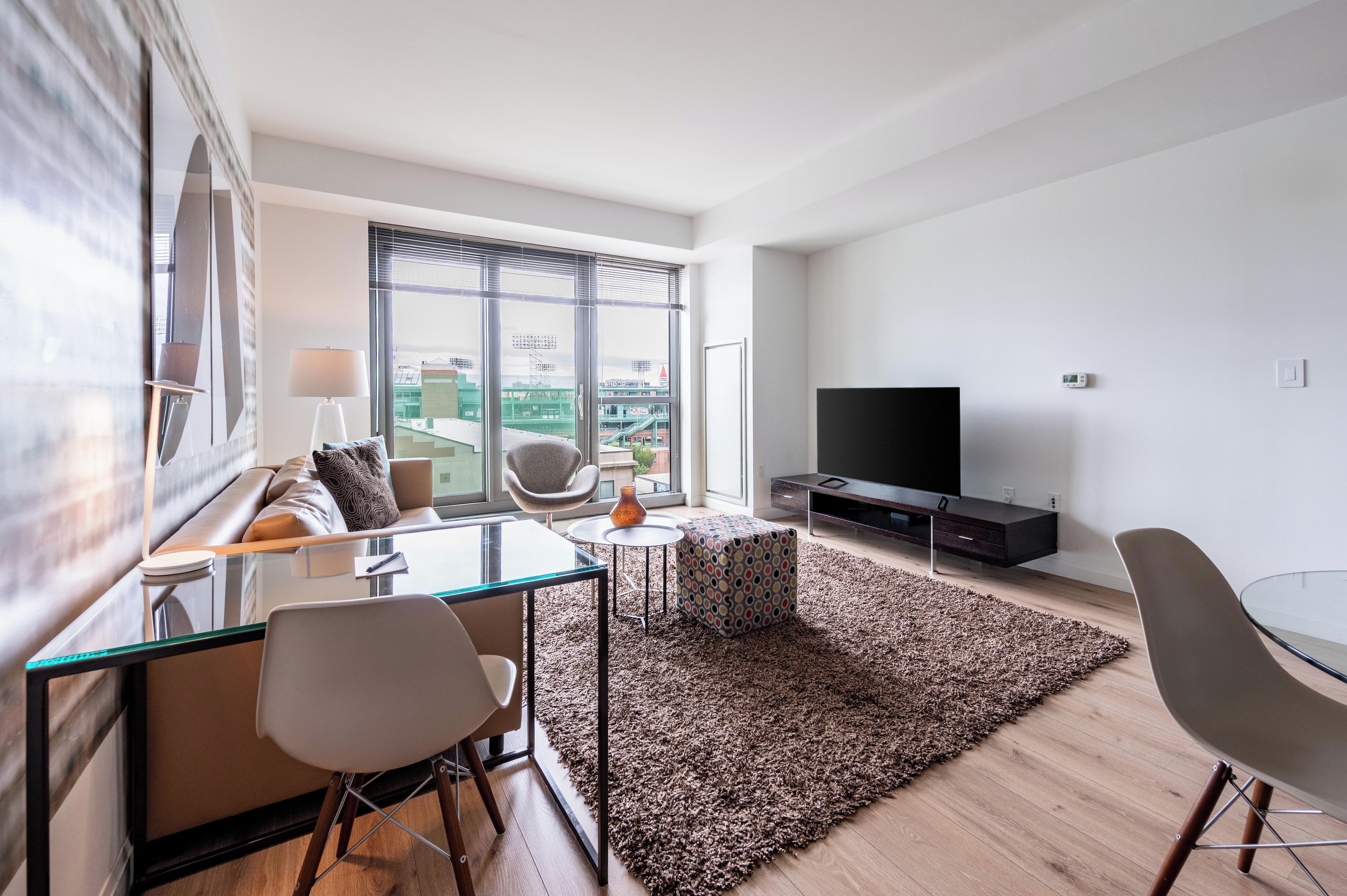 Een moderne woonkamer met een glazen eettafel, een bank, een televisie en grote ramen met uitzicht op de stad.