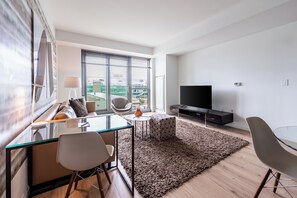 A modern living room with a glass dining table, a sofa, a TV, and large windows offering a city view.