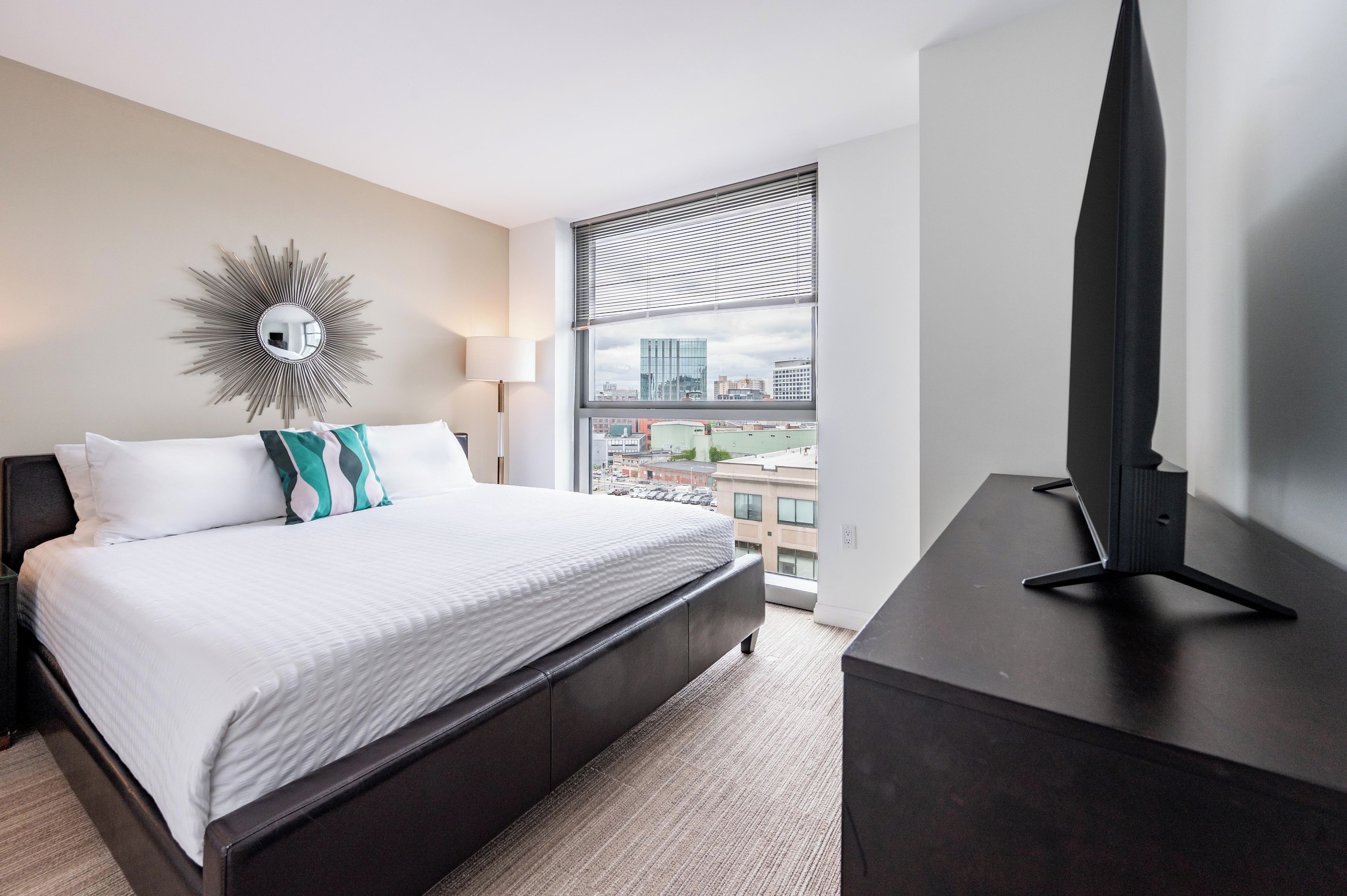A hotel room with a bed, a large window with blinds, a TV on a stand, and a decorative mirror on the wall.