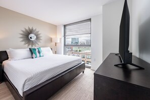 Habitación de hotel con cama, un ventanal con persianas, un televisor sobre un mueble y un espejo decorativo en la pared.
