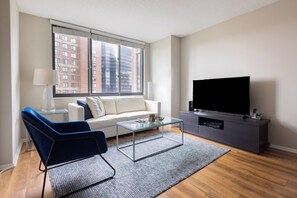 A modern living room with a sofa, a glass coffee table, a television, and a large window with a city view. - Gorgeous One Bedroom - 5-min Walk from Metro-North (White Plains)