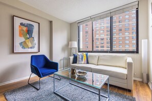A modern living room with a glass coffee table, a blue chair, a white sofa, and a large window with a city view.