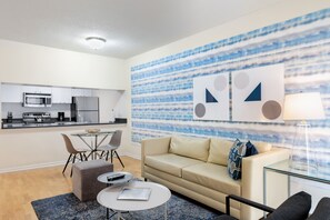 Un salon moderne avec un canapé, une table basse et des fauteuils. On aperçoit une cuisine équipée à l’arrière-plan.