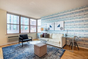 A modern living room with a large window, a beige sofa, a black armchair, a round coffee table, and a blue patterned rug.