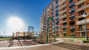 Un parque infantil con juegos, un sendero de ladrillos y un edificio residencial de varios pisos con balcones.