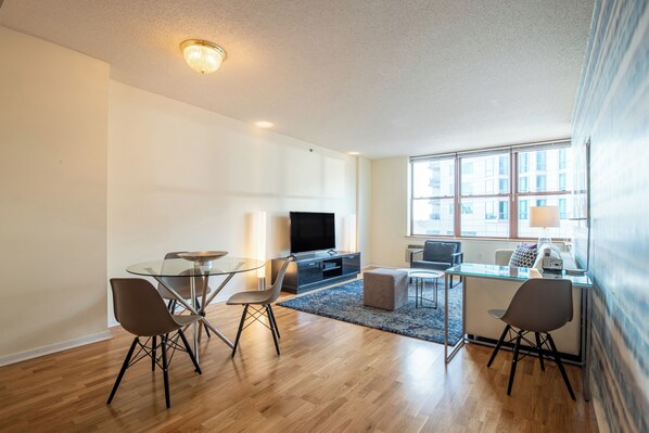 A modern living room with a glass dining table, two chairs, a sofa, a TV, and a large window.