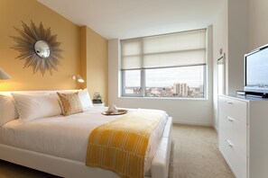 Quarto de hotel com cama, uma janela grande com persianas, televisão e um espelho decorativo na parede.