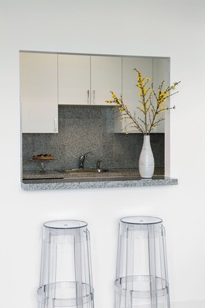 Una cocina con armarios blancos, encimera de granito, un florero con flores amarillas y dos taburetes de vidrio.